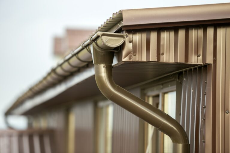 Vue d'un système de gouttière et de descente d'eau en métal marron sur le bord d'un toit en tôle ondulée, conçu pour éloigner l'eau de pluie du bâtiment.