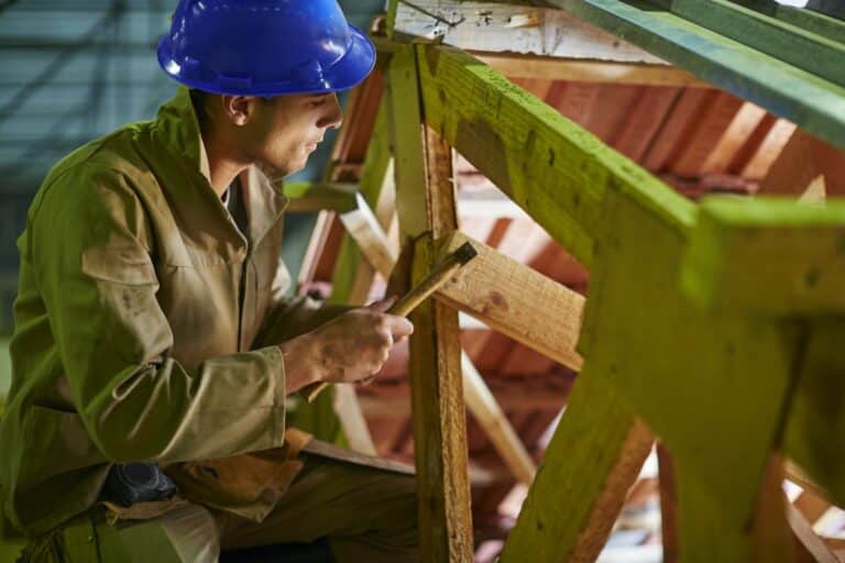 Un ouvrier du bâtiment portant un casque bleu et des vêtements de travail marron utilise un marteau sur une structure en bois sur un chantier de construction.