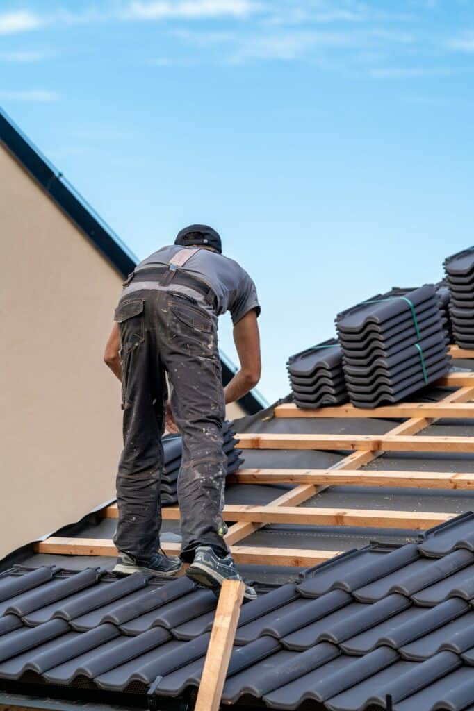 Un ouvrier du bâtiment installe des tuiles sur un toit en pente. Une pile de tuiles est posée sur la charpente en bois à côté de lui. Le ciel est clair en arrière-plan.