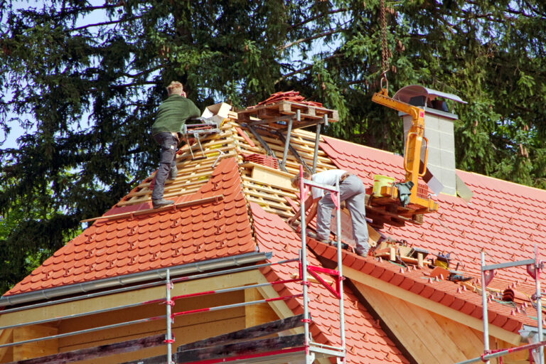 Deux ouvriers sont sur un toit à Figeac, en train de poser des tuiles rouges. Des échafaudages sont installés autour du bâtiment et des matériaux de construction sont dispersés à proximité. Des arbres sont visibles en arrière-plan. Cette couverture artisanale à Figeac garantit un savoir-faire de qualité et un souci du détail.