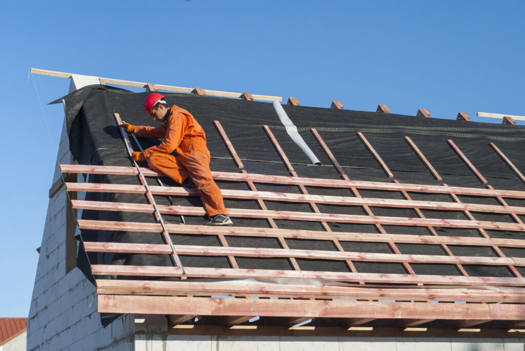 Un ouvrier du bâtiment en tenue orange et casque rouge travaille sur un toit, fixant des poutres en bois contre un ciel bleu clair. Cette scène capture parfaitement l'essence de la couverture à Puy-l'Évêque, mettant en valeur un savoir-faire artisanal dans des conditions sereines.
