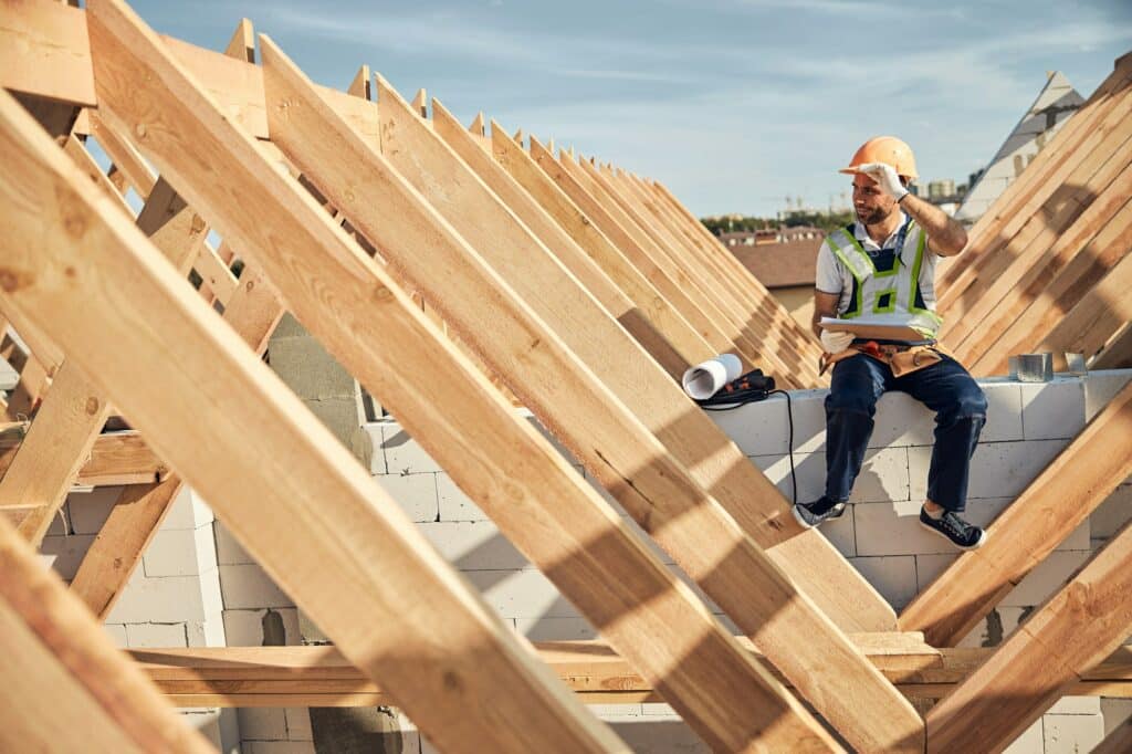 Un ouvrier du bâtiment portant un gilet réfléchissant et un casque est assis sur une charpente de toit, tenant un plan, par une journée ensoleillée.