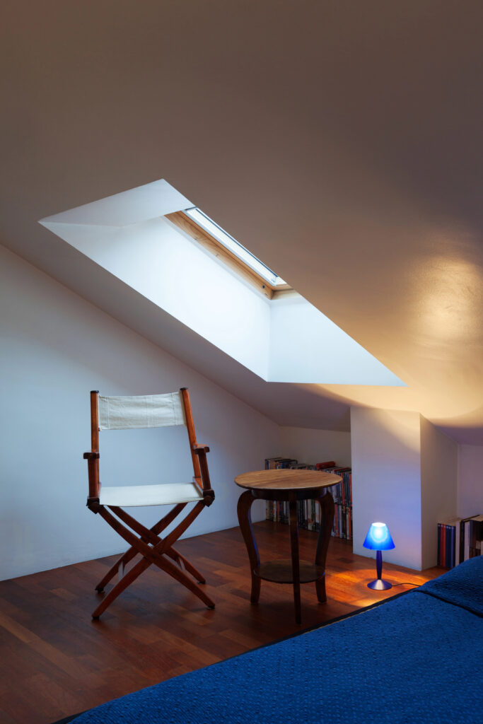 Une chambre minimaliste au plafond incliné comprend une lucarne de la récente pose de velux à Souillac, une chaise de réalisateur en bois, une petite table en bois, une lampe bleue et un lit recouvert d'une couverture bleue.