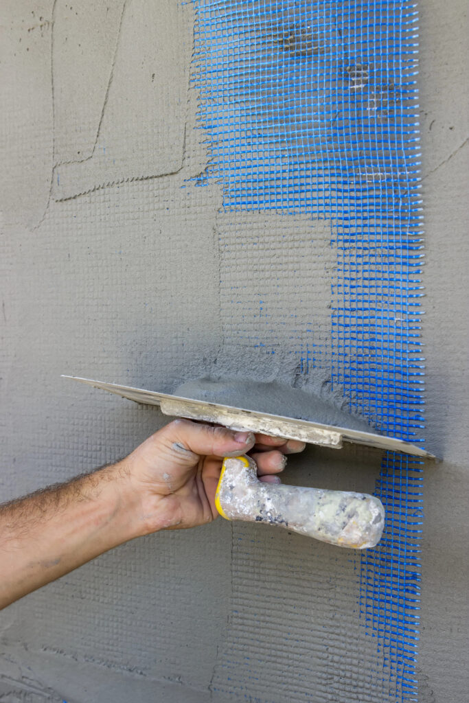Une main tenant une truelle applique du ciment sur du ruban adhésif sur un mur, rappelant les techniques expertes de ravalement de façade à Figeac.