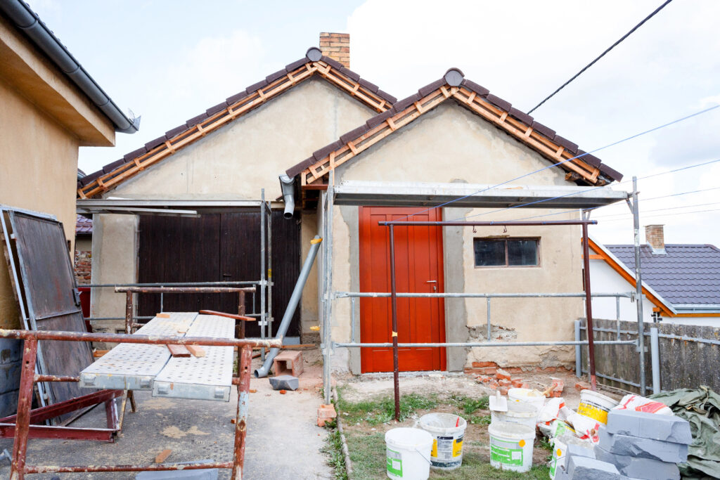 Une maison en rénovation avec des échafaudages, des matériaux de construction et des outils éparpillés un peu partout. Les murs extérieurs sont partiellement plâtrés, signe d'un ravalement de façade à Puy-l'Évêque, et une porte est peinte en rouge.