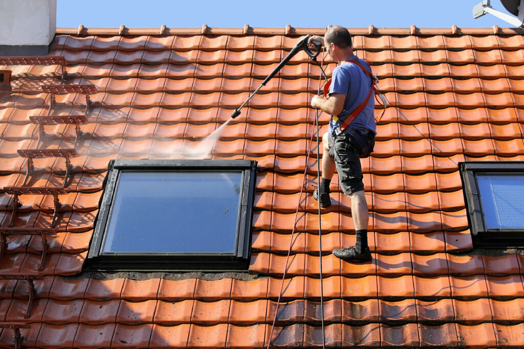 Un homme portant un harnais de sécurité lave sous pression un toit de tuiles près d'une fenêtre, effectuant un démoussage de toiture à Souillac.