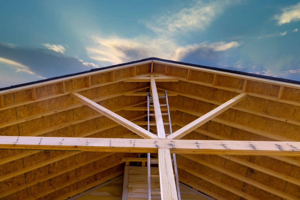 Vue de l'intérieur d'un toit en construction, montrant des poutres en bois et une échelle menant au sommet, avec un ciel bleu et des nuages visibles en arrière-plan.