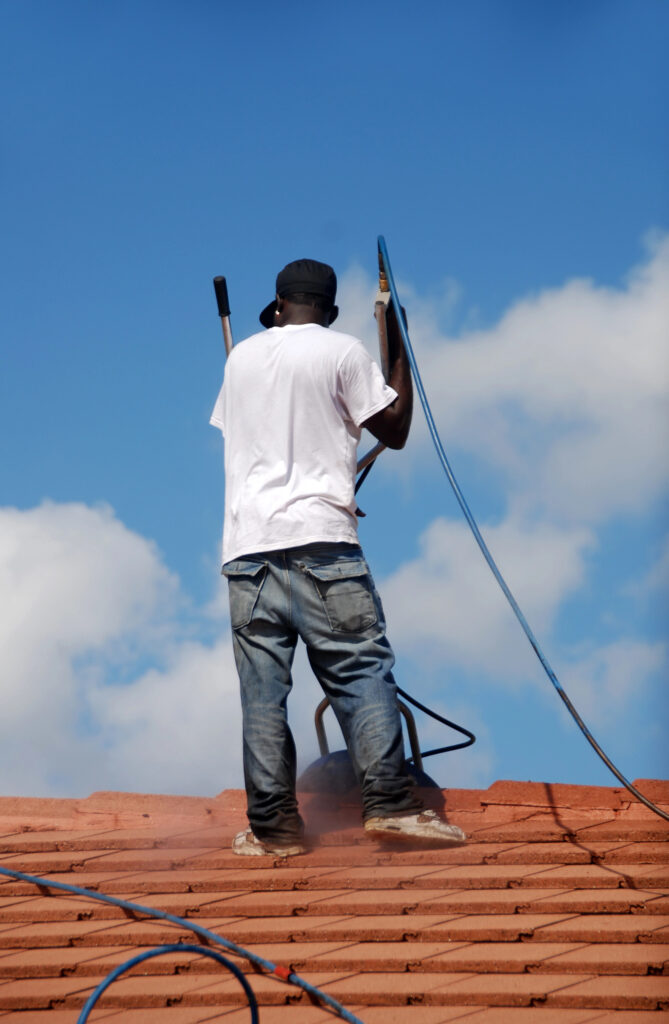 Une personne debout sur un toit, tenant un tuyau et du matériel, effectue un nettoyage de toiture à Gramat sur fond de ciel bleu avec quelques nuages.