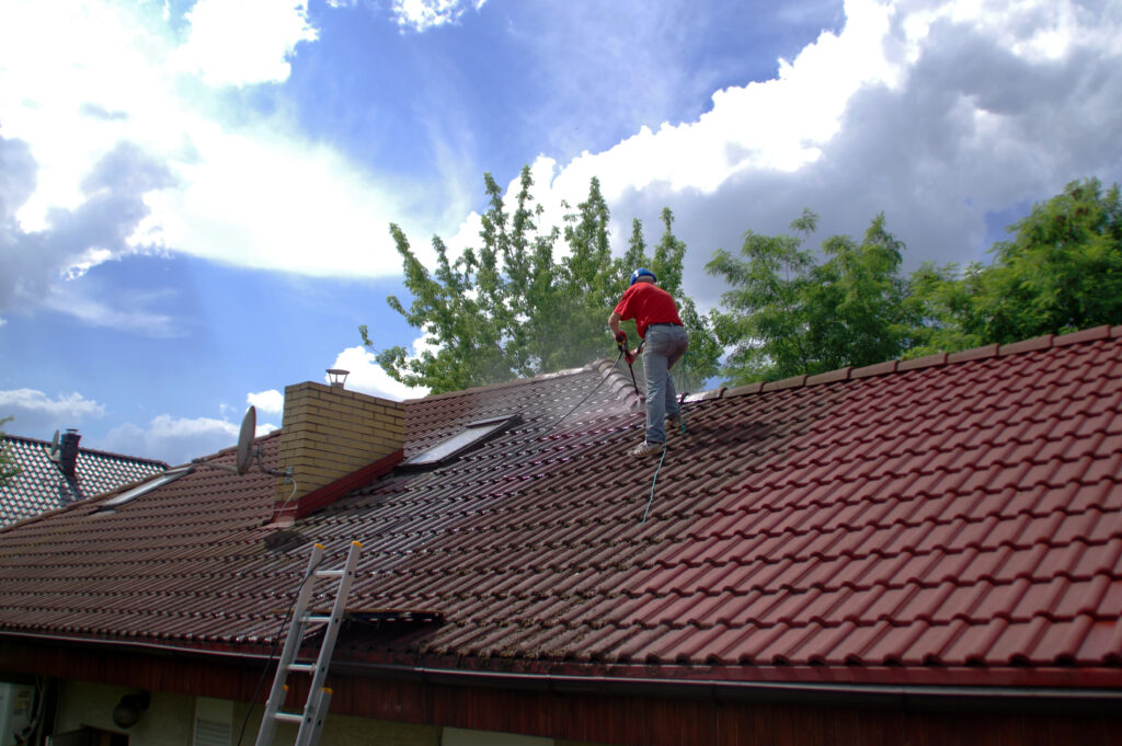 Une personne effectue un Nettoyage de toiture à Puy-l'Évêque, nettoyant les débris d'un toit de tuiles rouges à l'aide d'un nettoyeur haute pression. Une échelle est appuyée contre le toit et le ciel apparaît partiellement nuageux.