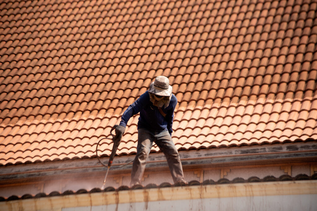 Une personne portant des vêtements de protection et tenant un pistolet travaille sur un toit en tuiles de terre cuite en plein soleil, effectuant un démoussage de toiture à Gourdon.