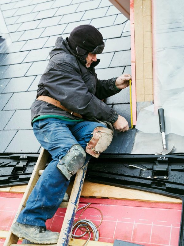 Un travailleur portant une veste et un équipement de sécurité est assis sur un toit et mesure les bardeaux avec un ruban à mesurer. Des outils et des matériaux de construction sont autour de lui.