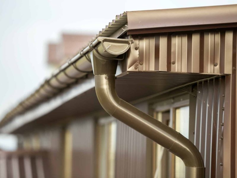 Vue d'un système de gouttière et de descente d'eau en métal marron sur le bord d'un toit en tôle ondulée, conçu pour éloigner l'eau de pluie du bâtiment.
