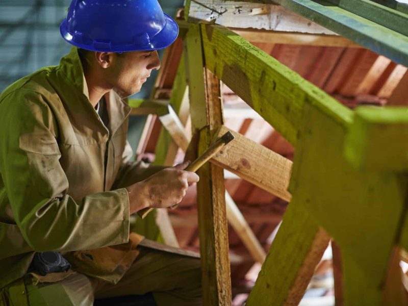 Un ouvrier du bâtiment portant un casque bleu et des vêtements de travail marron utilise un marteau sur une structure en bois sur un chantier de construction.