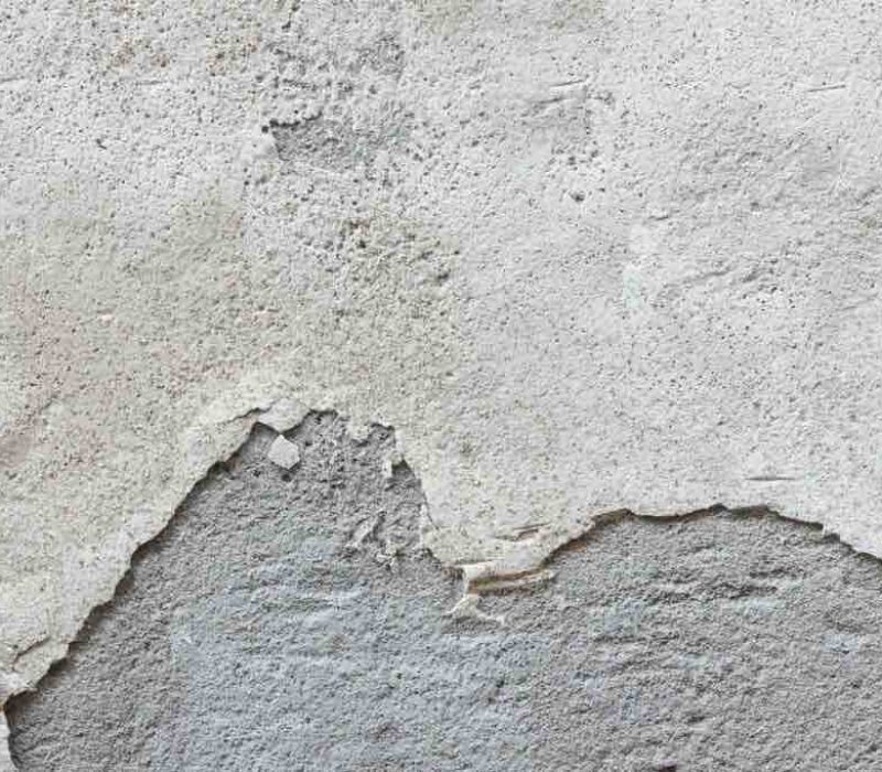 Vue rapprochée d'un mur texturé avec de la peinture beige écaillée révélant une surface grise en dessous. La peinture est craquelée et le mur est inégal.