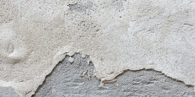 Gros plan d'un vieux mur texturé avec du plâtre écaillé, révélant une couche grise en dessous. La surface présente des signes d'usure et d'altération.