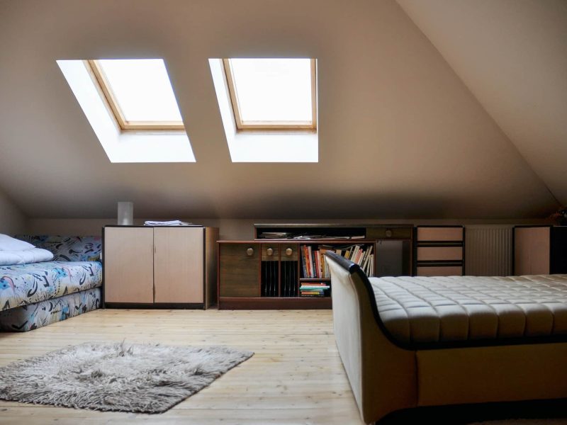Une chambre mansardée confortable avec un lit, un canapé, une bibliothèque et deux lucarnes au plafond incliné. La chambre dispose de parquet en bois de couleur claire et d'un petit tapis.