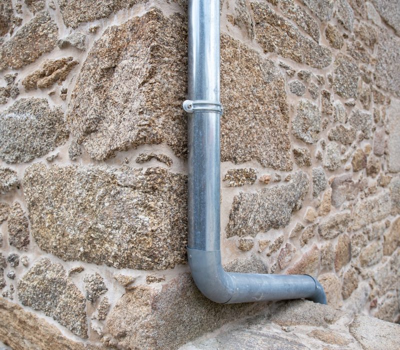 Un tuyau d'évacuation en métal fixé au coin de l'extérieur d'un bâtiment en pierre, éloignant l'eau de la structure.