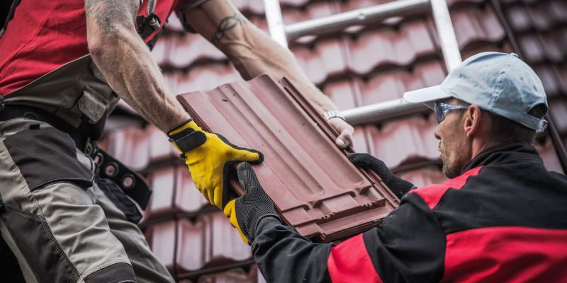 Deux ouvriers en uniforme manipulent des tuiles sur un toit avec une échelle métallique à proximité.