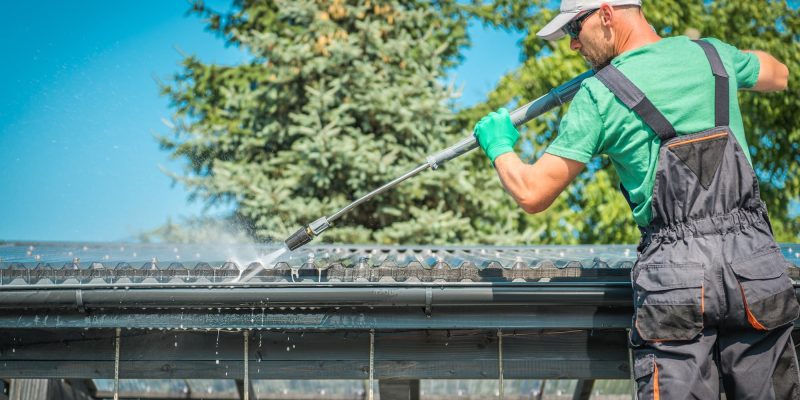 Un homme en combinaison de travail et gants utilise un tuyau d'eau à haute pression pour nettoyer une gouttière de toit par une journée ensoleillée.