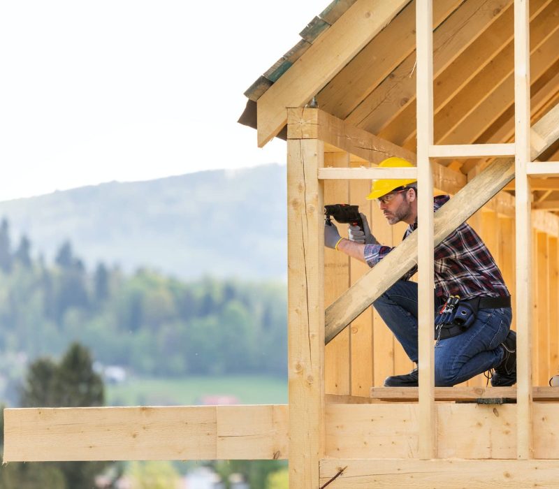 Un ouvrier du bâtiment portant un casque de sécurité utilise un outil électrique sur la charpente en bois d'une maison en construction. Des arbres et des collines sont visibles en arrière-plan.