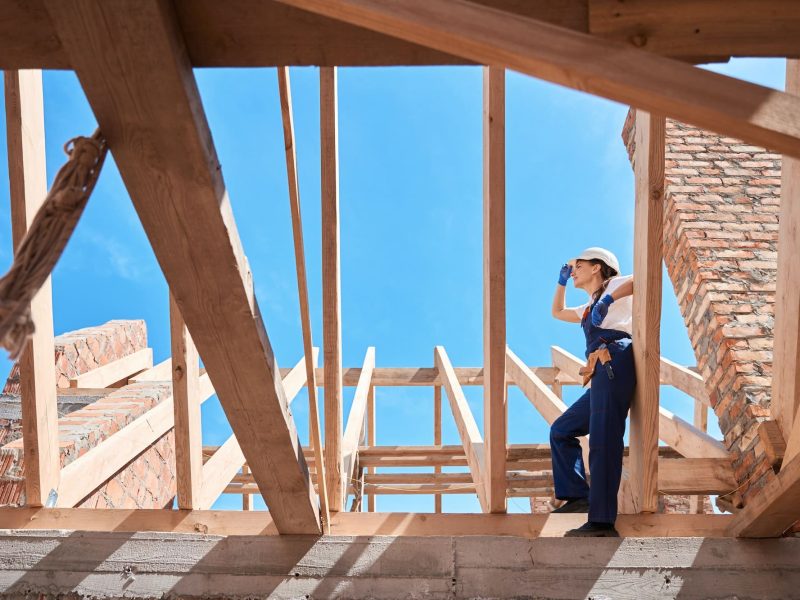 Un ouvrier du bâtiment portant un casque et une combinaison se tient debout sur les poutres en bois d'un toit inachevé, regardant au loin.